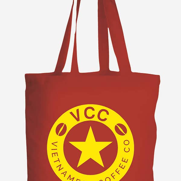 A red VCC branded tote bag. The bag is emblazoned with a large round yellow, circular Vietnamese Coffee Co. logo with a star at it's centre.